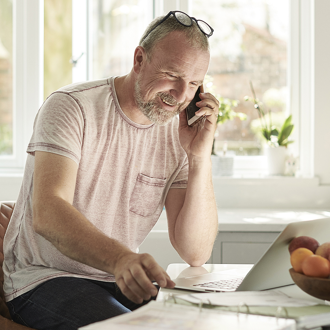 A man with glasses sitting atop his head sits in front of a laptop while talking on the phone