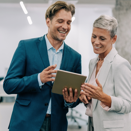 Man and woman stand, smiling as they look at tablet