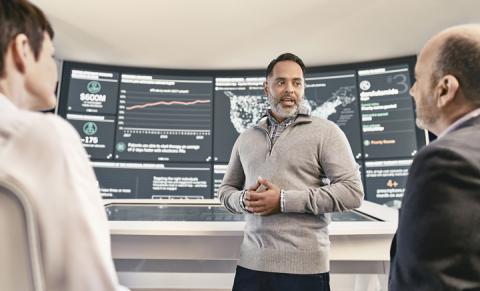 Man standing in front of large digital displays giving a presentation to two people.