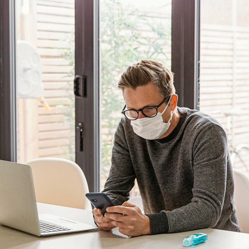 Man in front of laptop looks at phone while wearing a mask