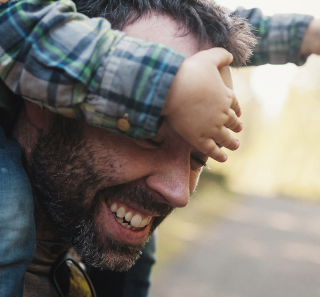 Father smiles. His eyes are covered as he carries child on shoulders