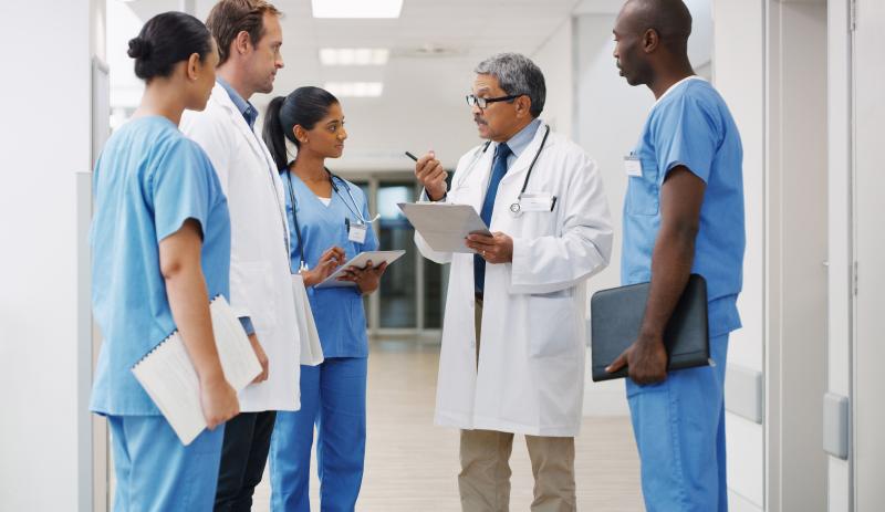 Doctor stands and talk with four other medical professionals in middle of hospital hallway