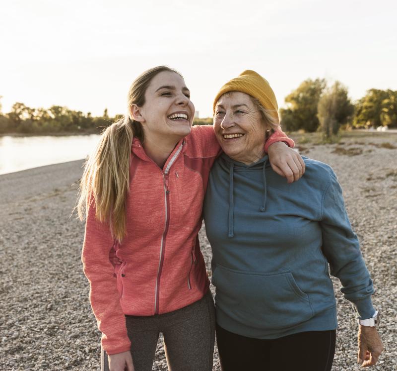 Older and younger adult women embracing, smiling, and walking along a lake shore.