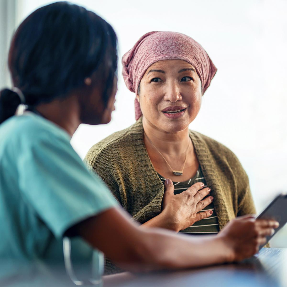 A medical professional who is wearing scrubs holds a tablet and speaks with a patient